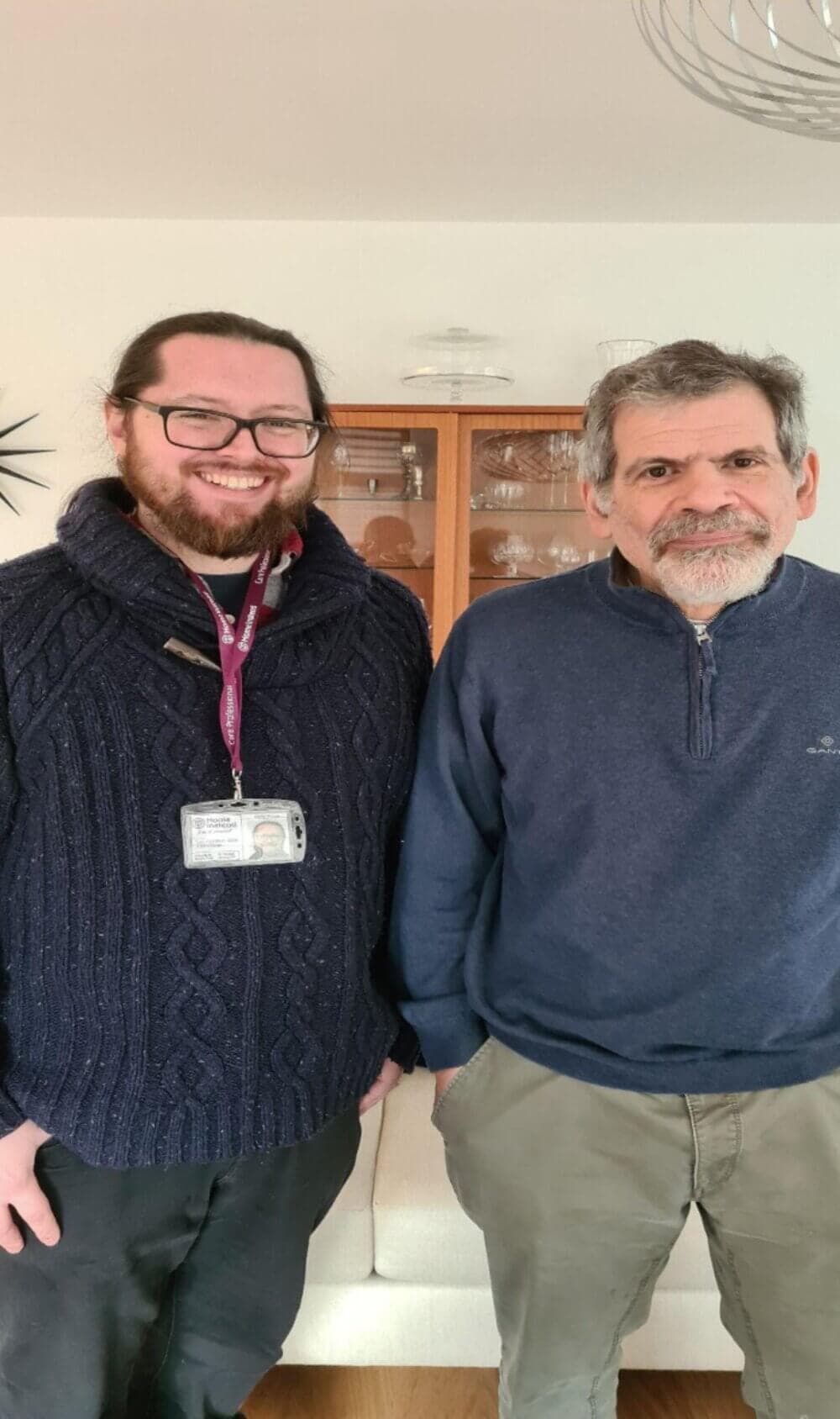 Two men are standing side by side, smiling, indoors. One wears glasses and an ID badge, the other has a beard and mustache. - Home Instead