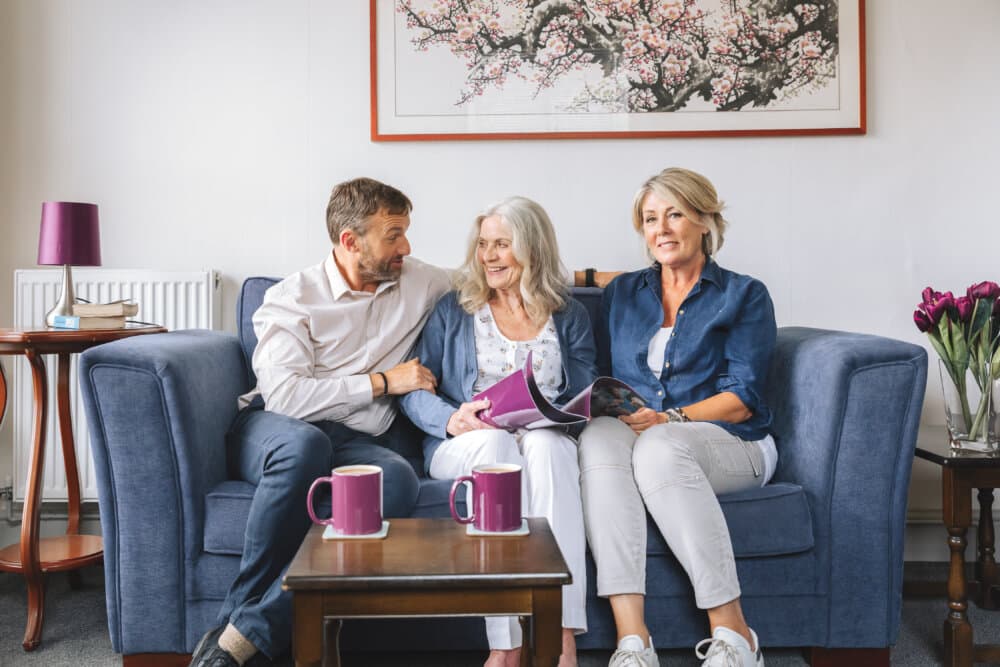 Three smiling people sitting on a blue couch, sharing a photo album, with two purple mugs and a framed painting behind them. - Home Instead