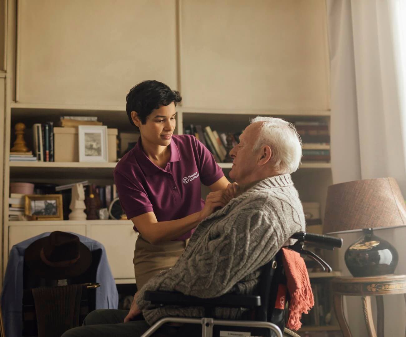 A caregiver helps an elderly man in a wheelchair with his sweater in a cozy, book-filled room. - Home Instead