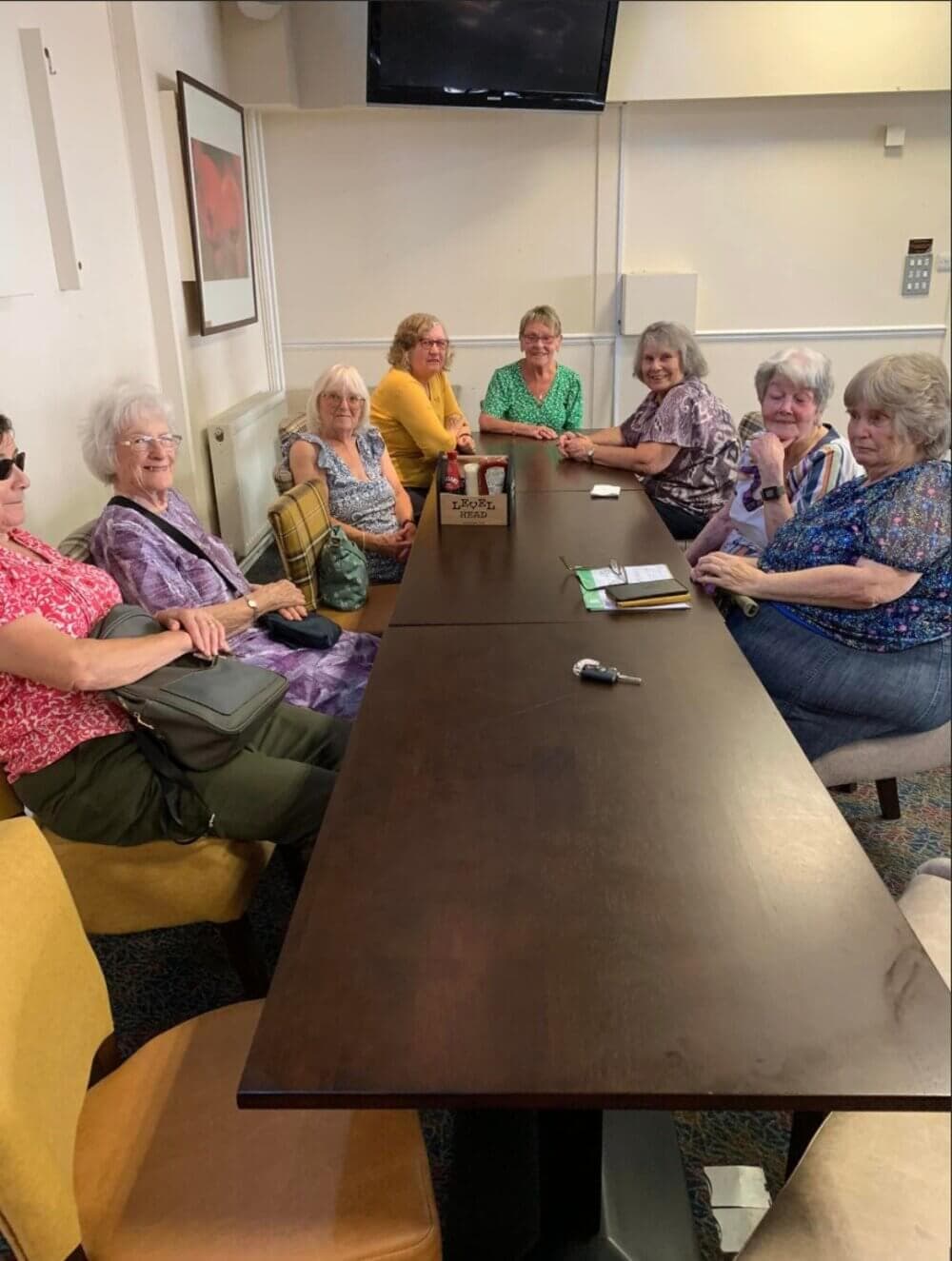 A group of older women sitting around a long table in a restaurant, smiling at the camera. - Home Instead