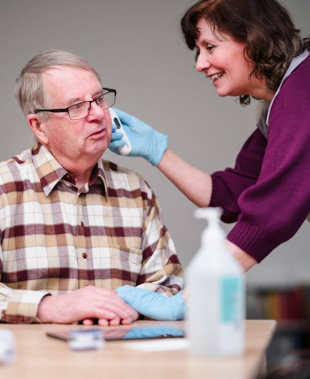 Healthcare worker helps an elderly man with ear care at a table, both smiling, with medical supplies nearby. - Home Instead