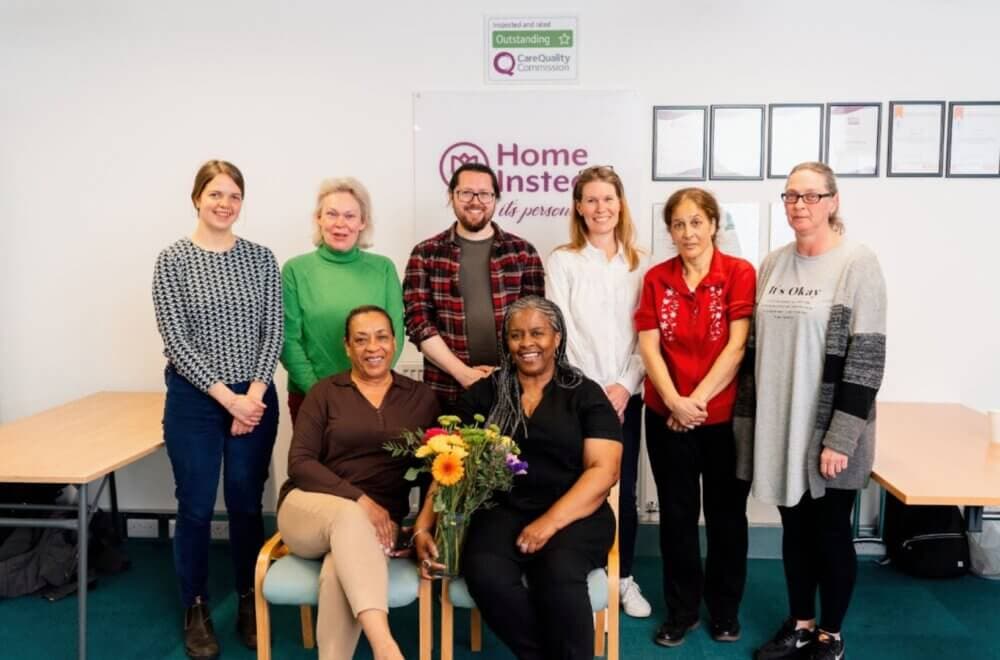 Group of eight people, some seated and some standing, smiling at the camera. A woman in front holds a bouquet of flowers. - Home Instead