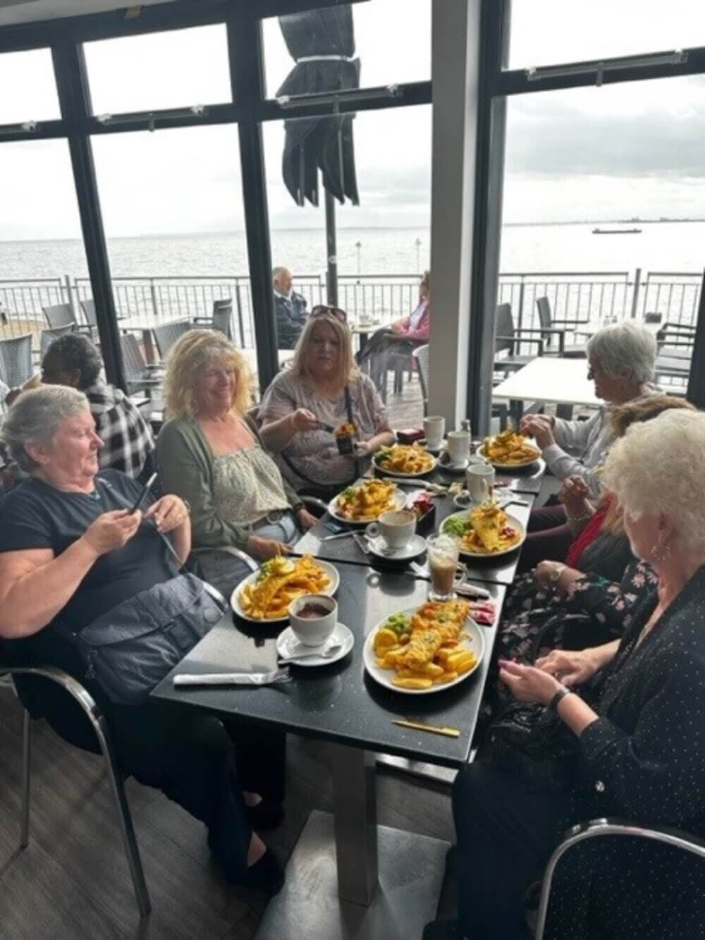 A group of women enjoying a meal together at a restaurant with large windows overlooking the water. - Home Instead