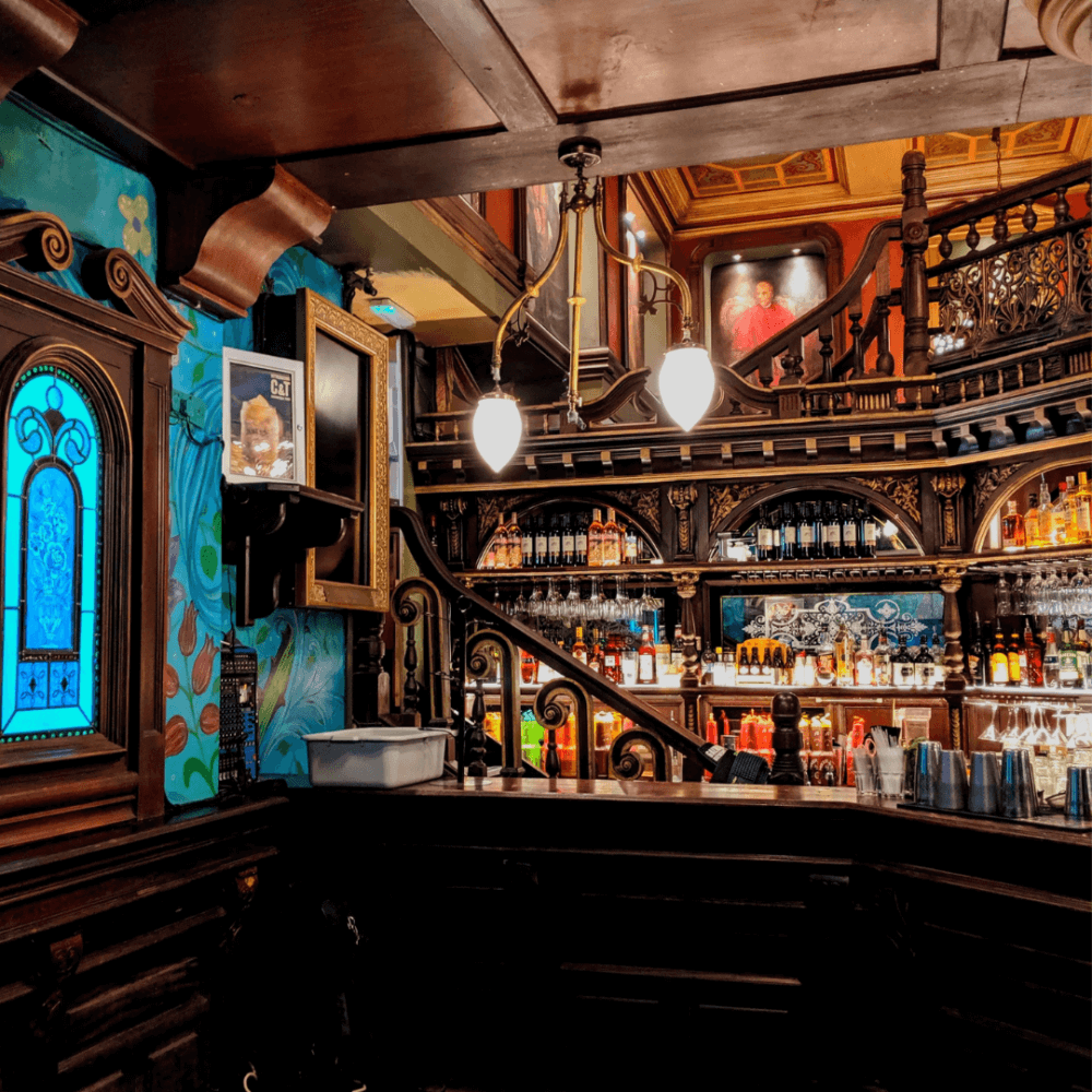 A cozy, ornate bar with wooden decor, a colorful blue stained glass window, and shelves filled with various bottles. - Home Instead