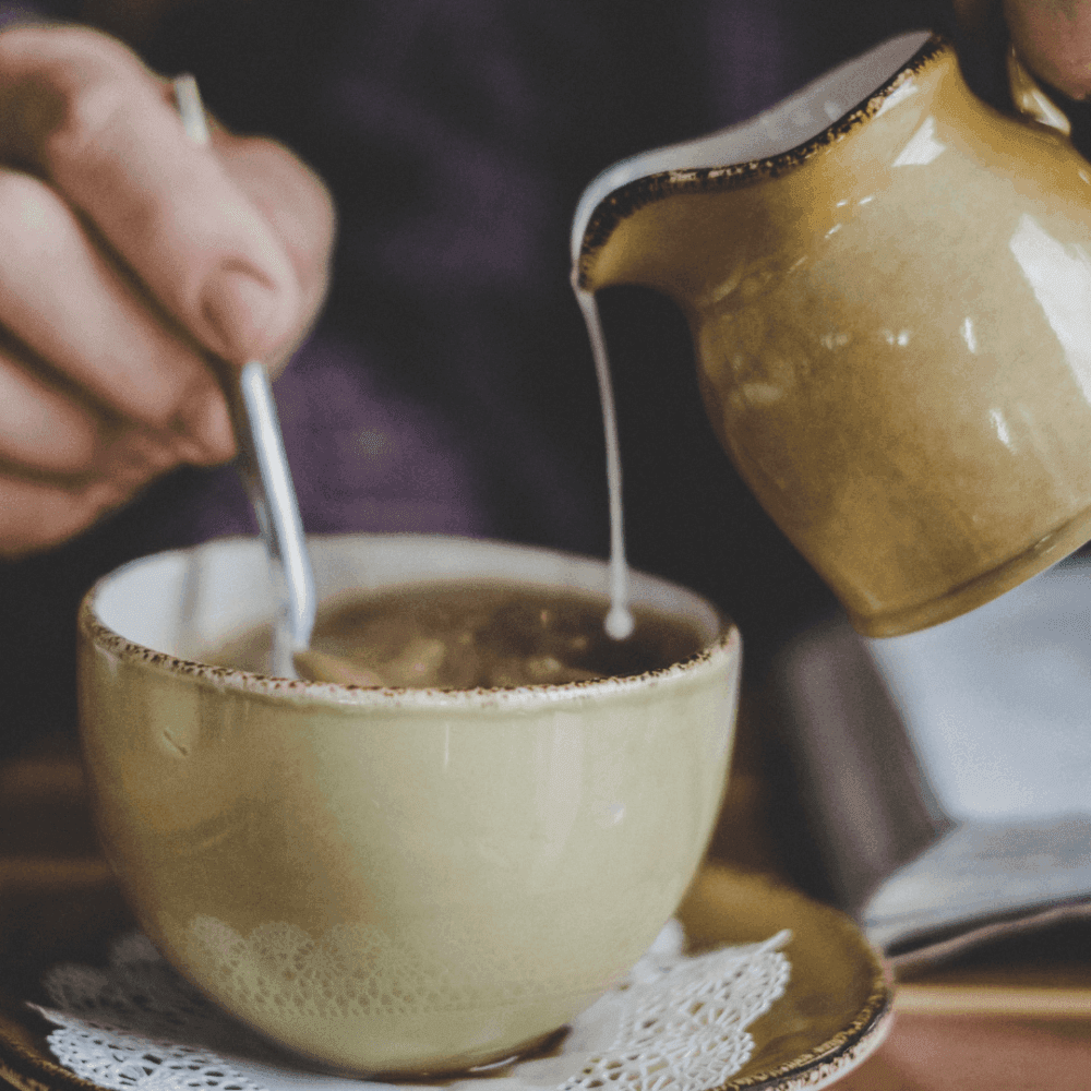 A person stirring a cup of coffee with a spoon, while holding a cream pitcher above the cup. - Home Instead
