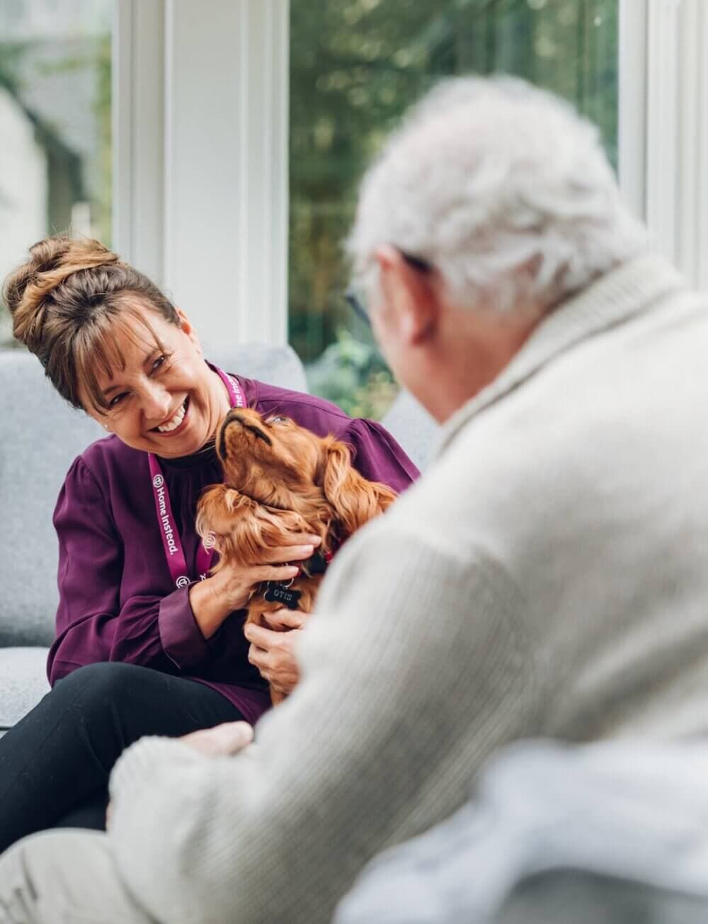 A woman smiles and holds a dog while sitting next to an elderly person in a cozy indoor setting. - Home Instead