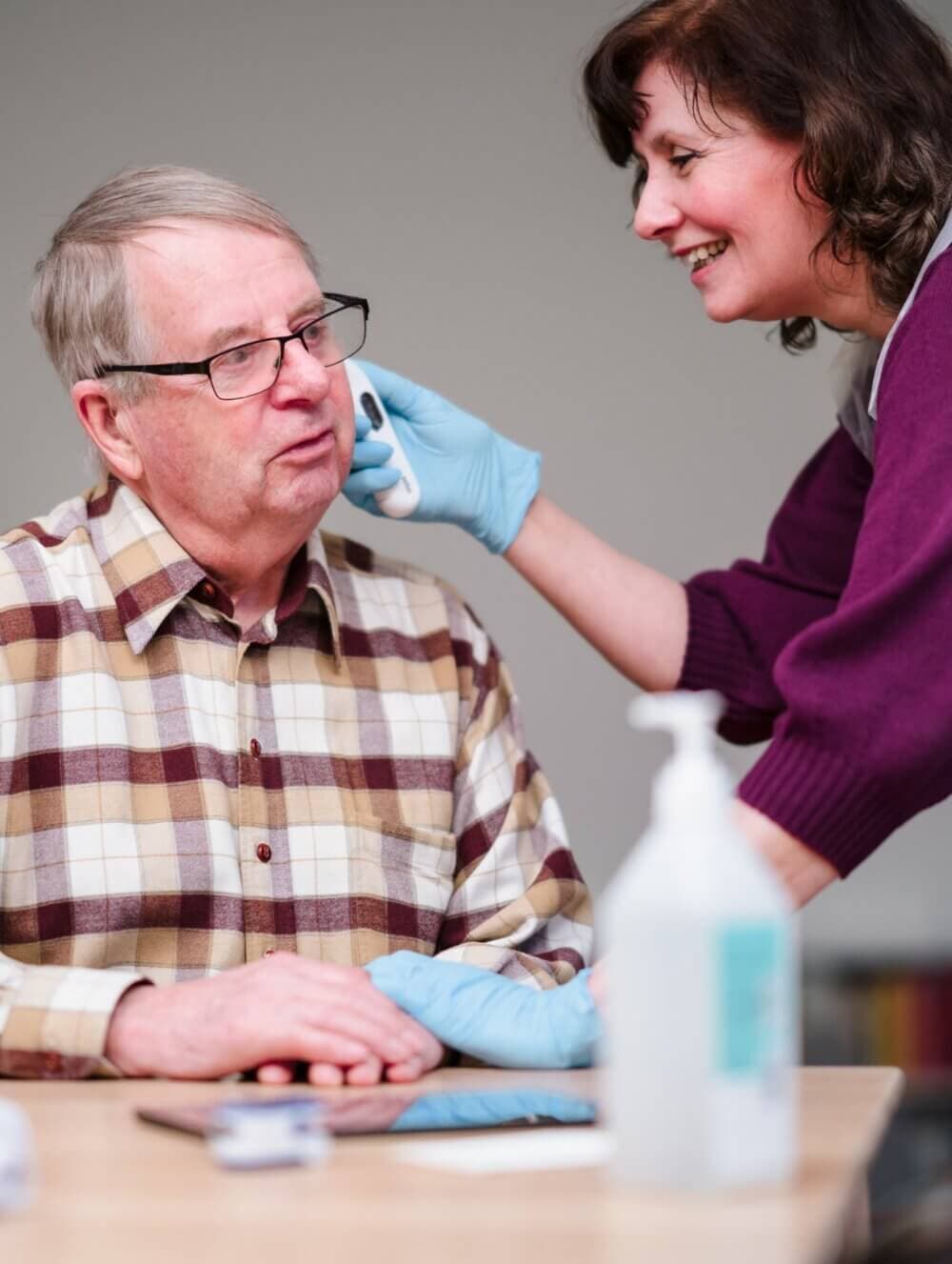 A caregiver in gloves smiles while assisting an elderly man, who is seated, with cleaning his ear. - Home Instead