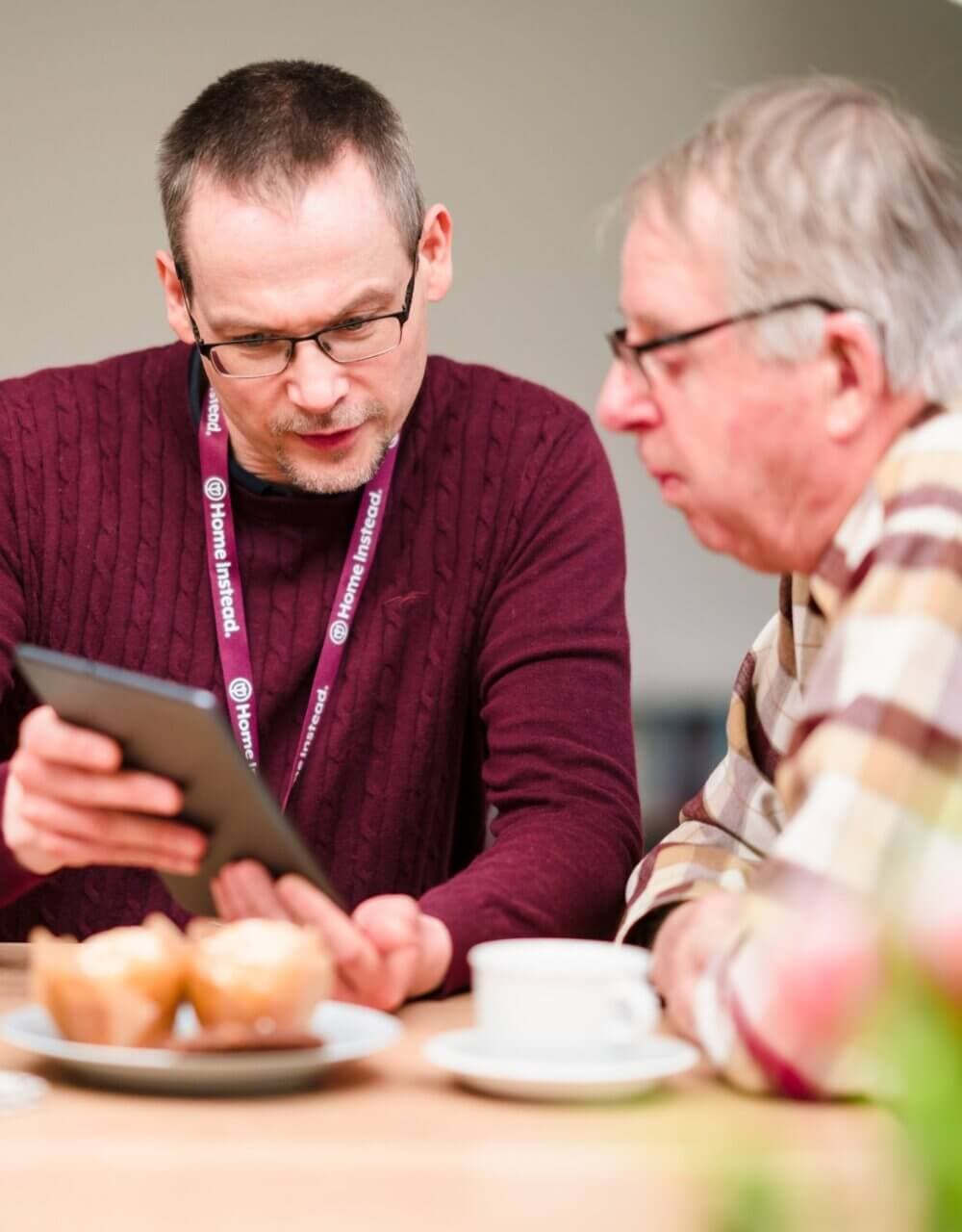 Two men looking at a tablet screen at a table with muffins and a cup, one wearing a name tag. - Home Instead
