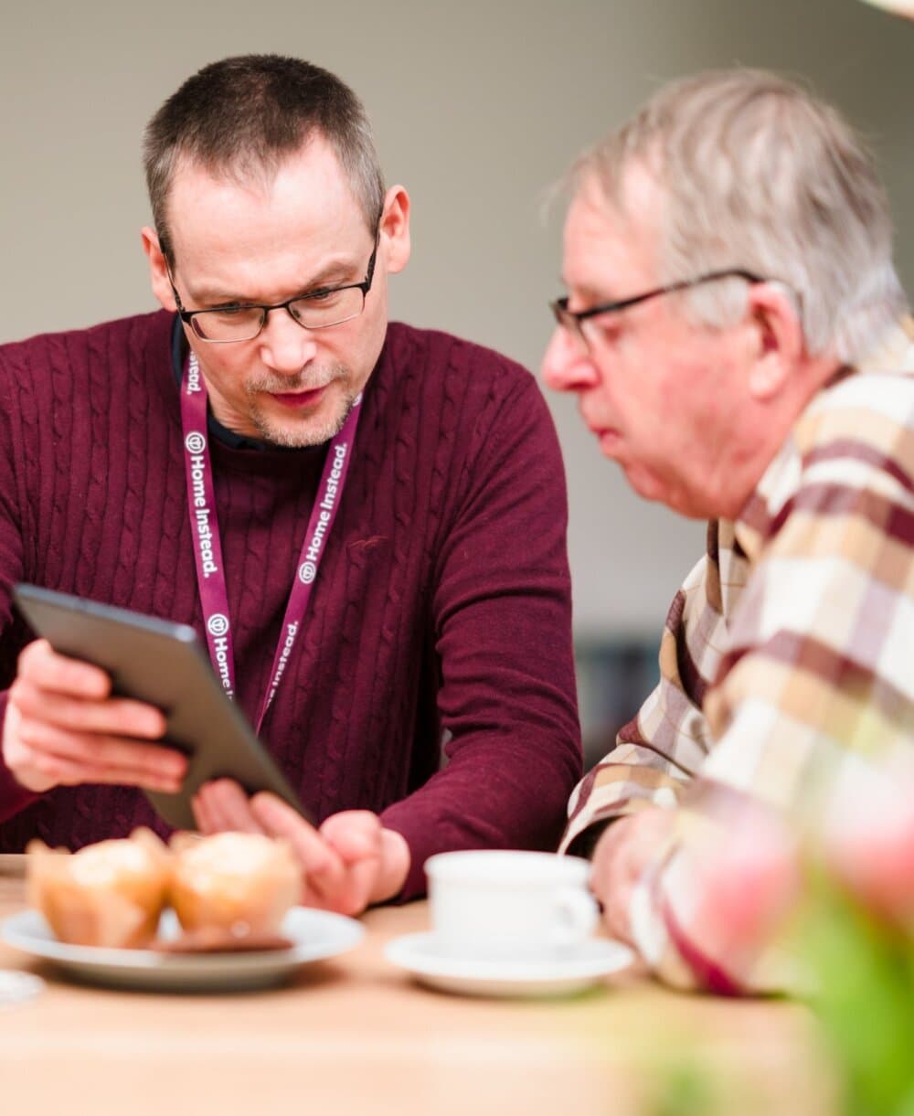 Two men sitting at a table with muffins and coffee, one showing the other something on a tablet screen. - Home Instead