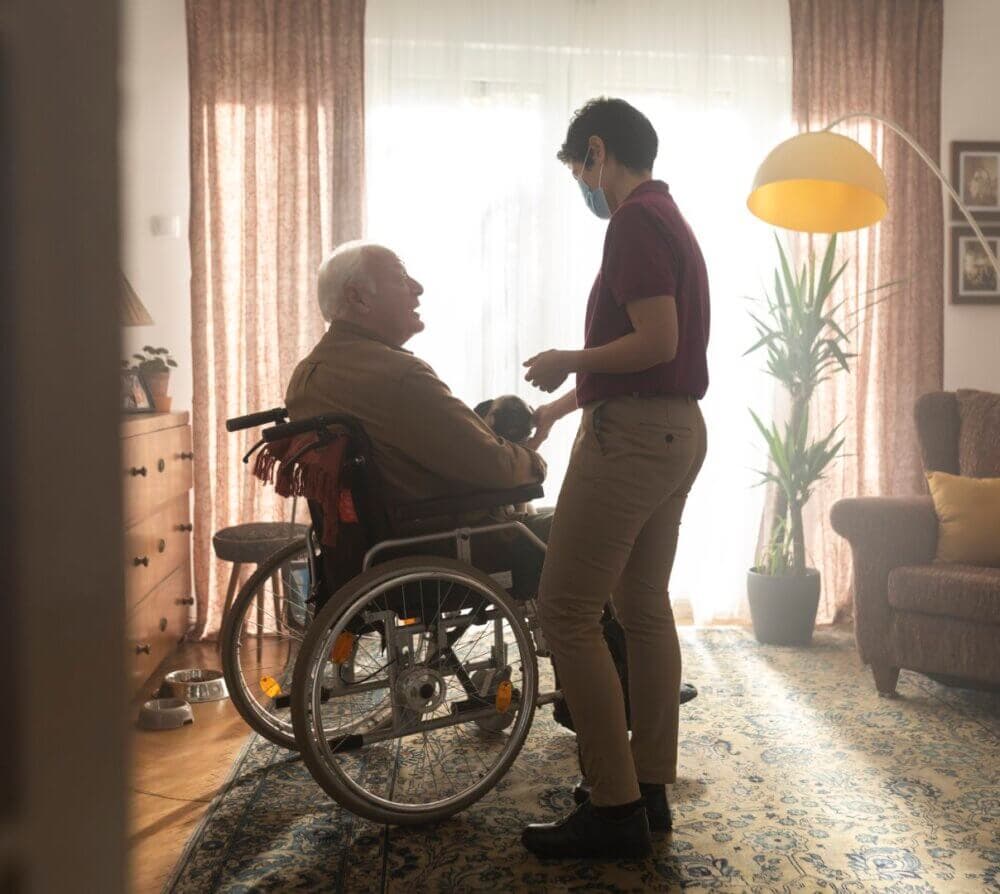 A caregiver wearing a mask tends to an elderly man in a wheelchair in a warmly lit living room. - Home Instead