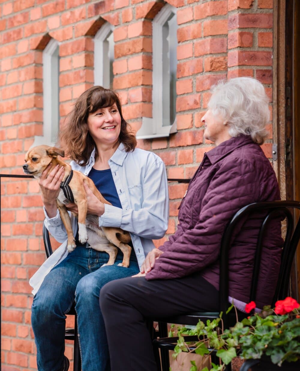 Two women sit on chairs outdoors by a brick wall, one holding a dog and smiling at the other. - Home Instead