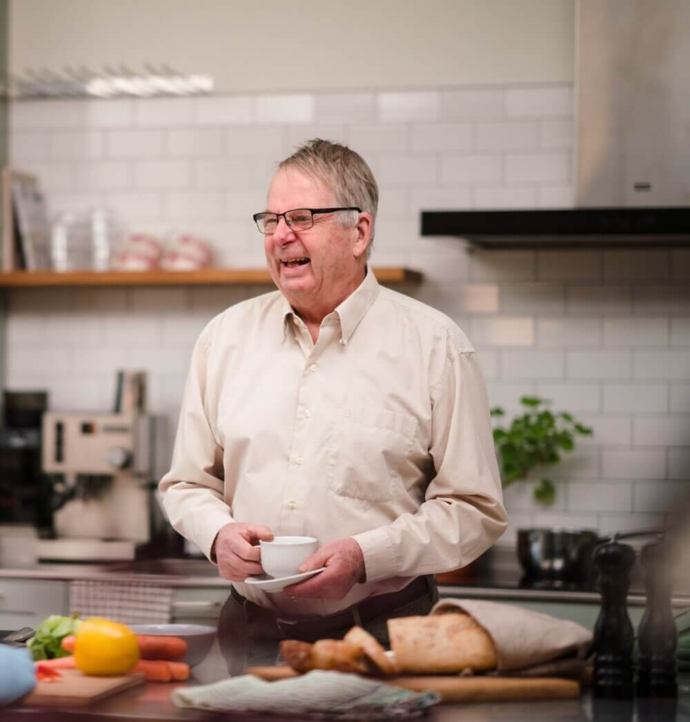 Smiling man holding a cup while standing in a kitchen with various foods on the counter. - Home Instead