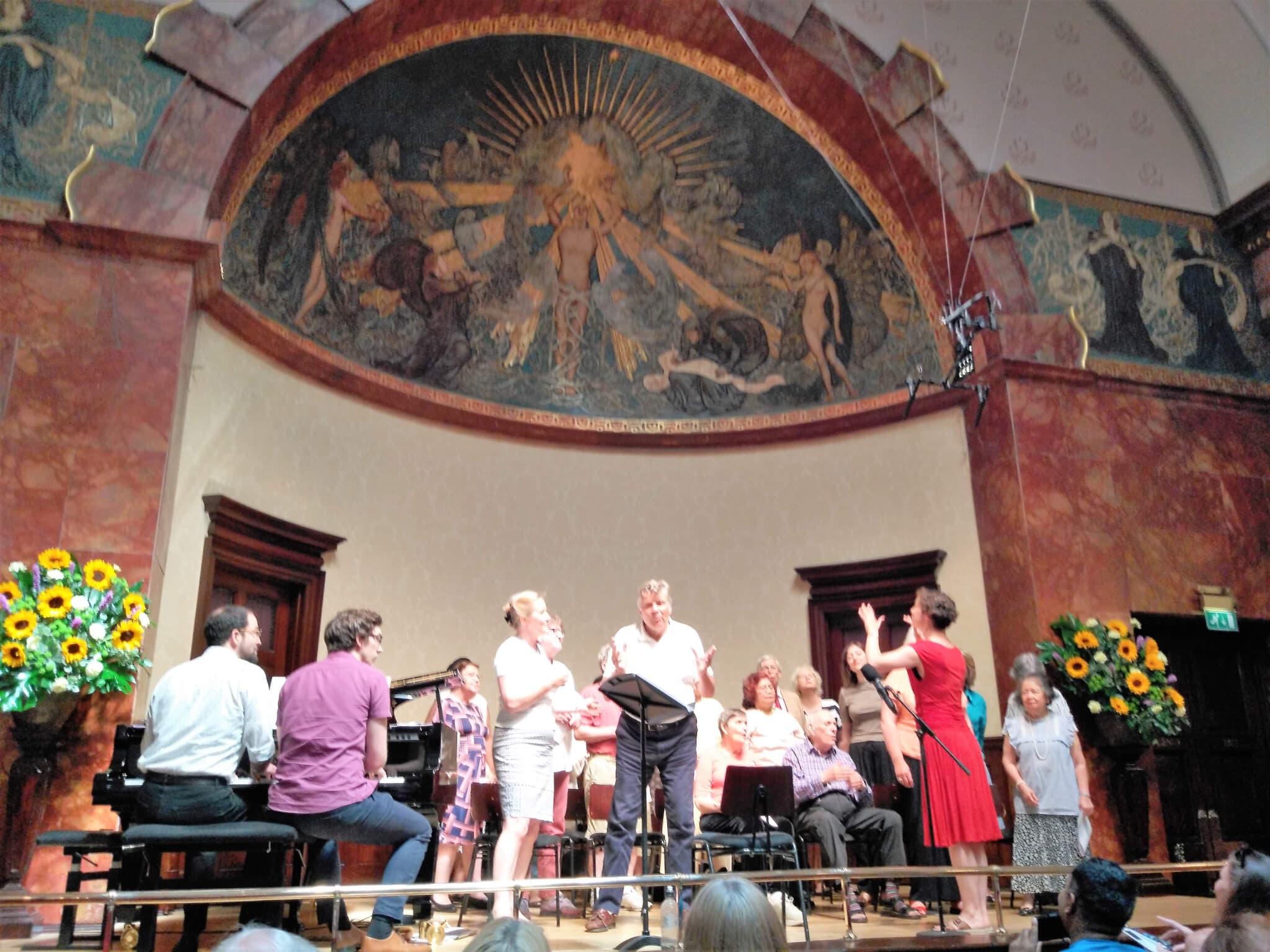 Group of singers performing on stage with a conductor and pianist, beneath a large, artistic mural. - Home Instead
