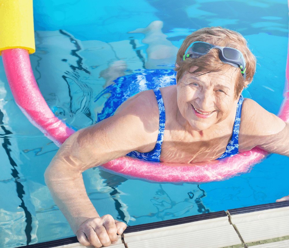 Elderly woman smiling while floating in a swimming pool with a pink pool noodle around her waist. - Home Instead