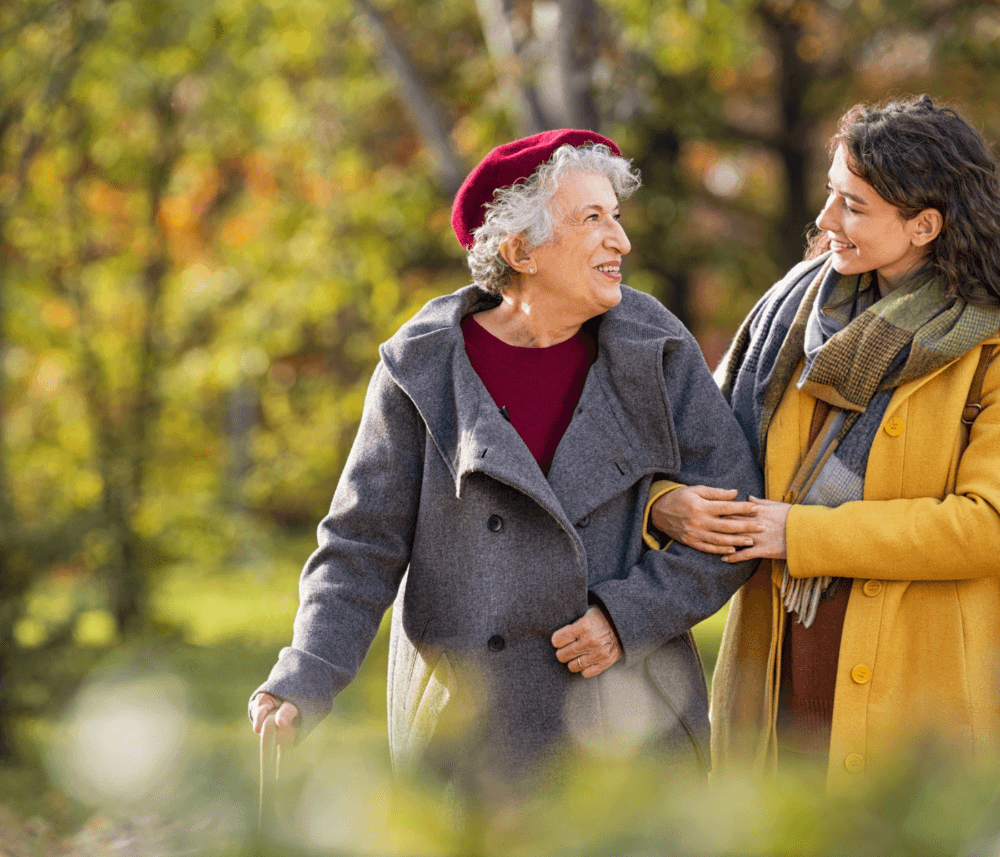 Elderly woman in grey coat and red beret walking with younger woman in yellow coat, smiling at each other outdoors. - Home Instead