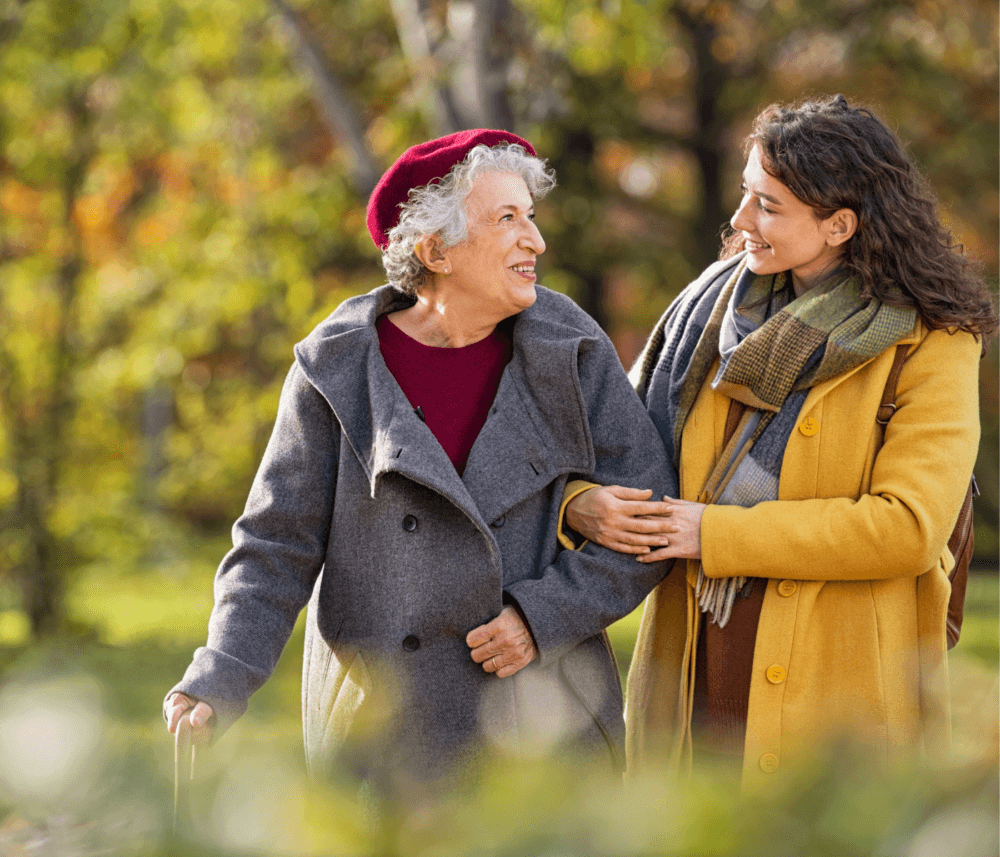 An older woman and a younger woman smile at each other while walking arm-in-arm through a park. - Home Instead