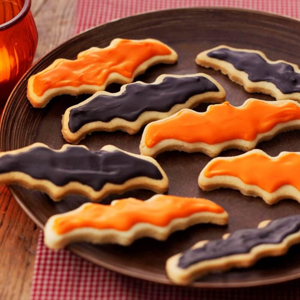 Plate of bat-shaped cookies, decorated with black and orange icing, on a wooden table with a red checked cloth. - Home Instead