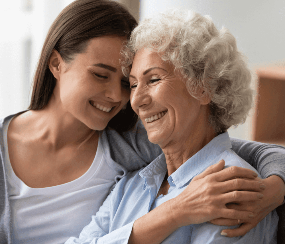 A young woman and an elderly woman share a warm embrace, smiling and leaning their heads together. - Home Instead