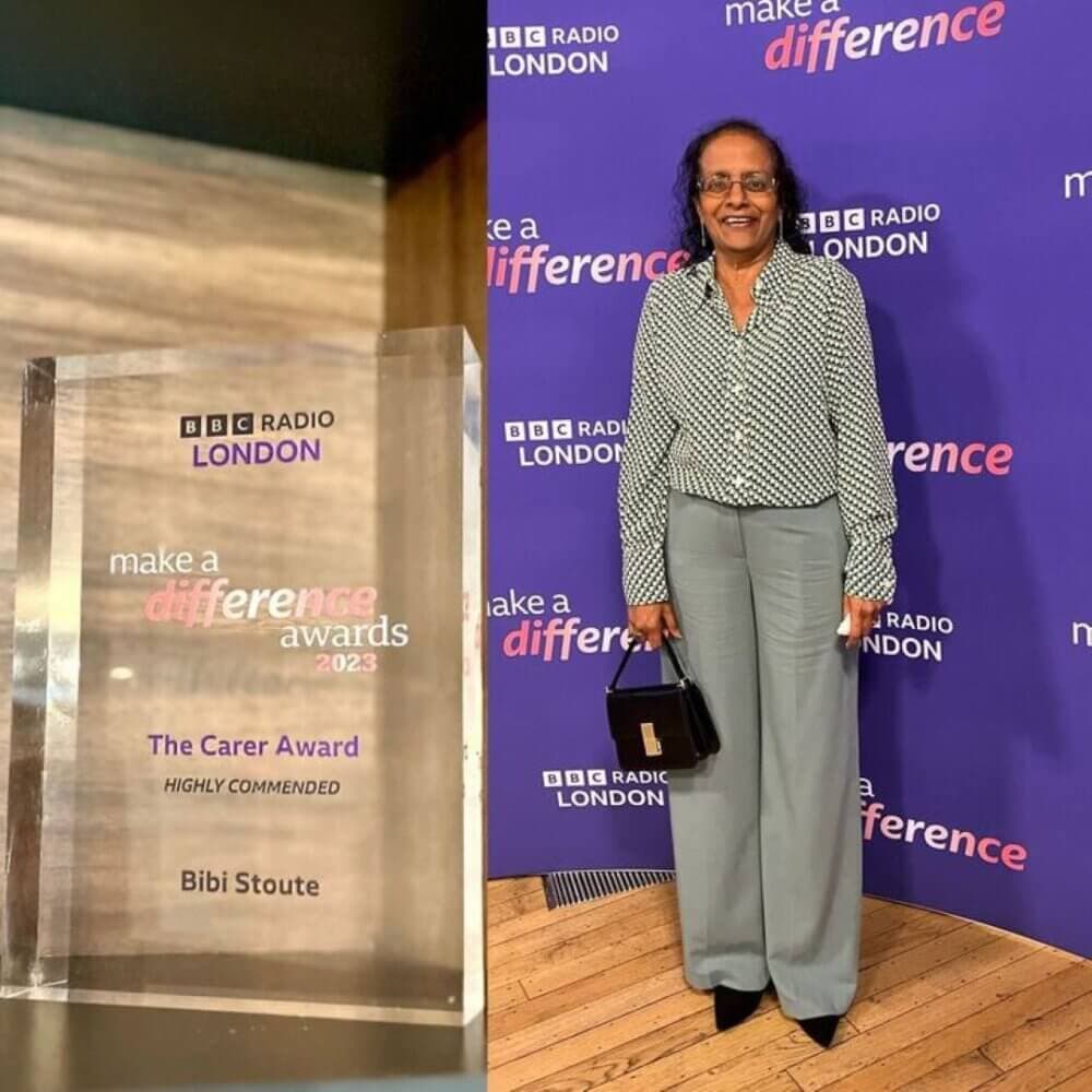 A person stands next to a BBC Radio London award display. The award reads "The Carer Award, Highly Commended, Bibi Stoute. - Home Instead