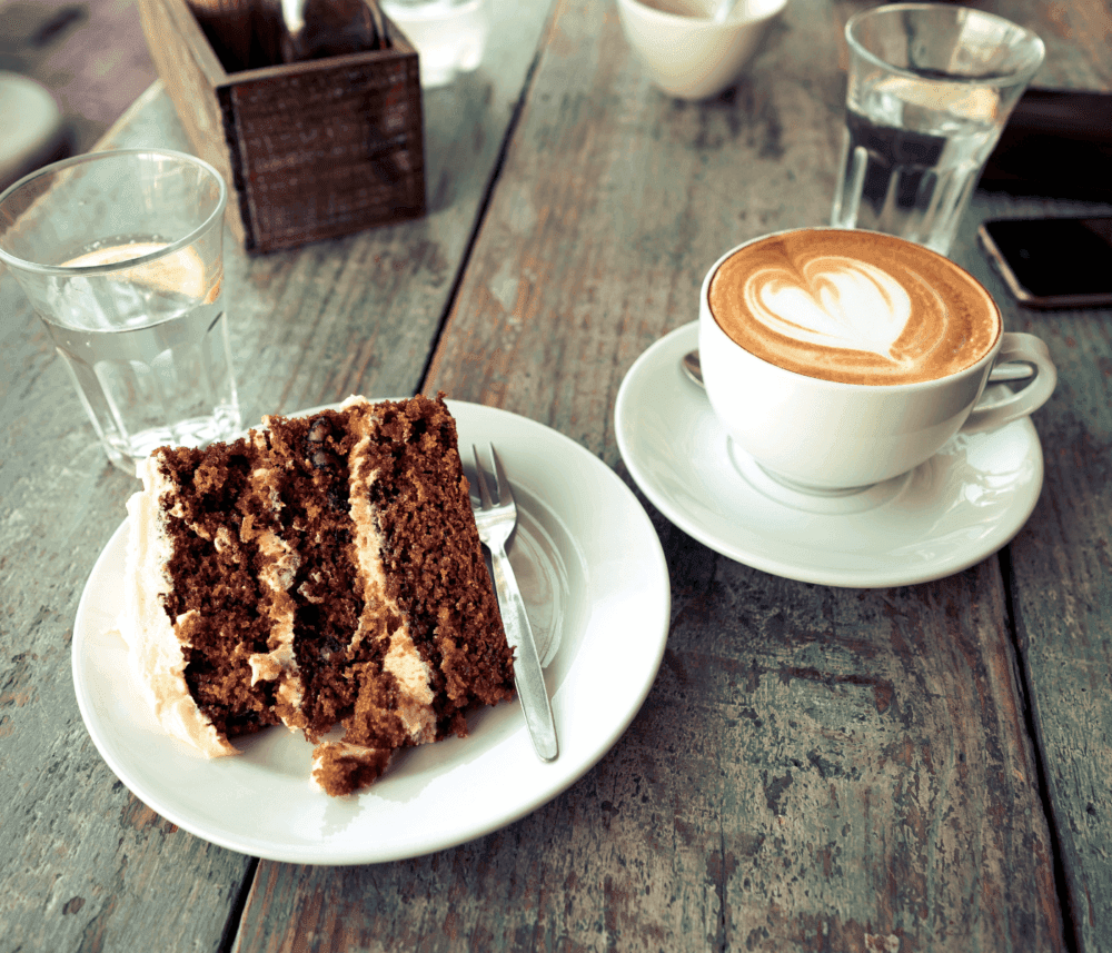 A slice of chocolate cake on a plate with a fork, next to a cup of latte with heart foam art and two glasses of water. - Home Instead