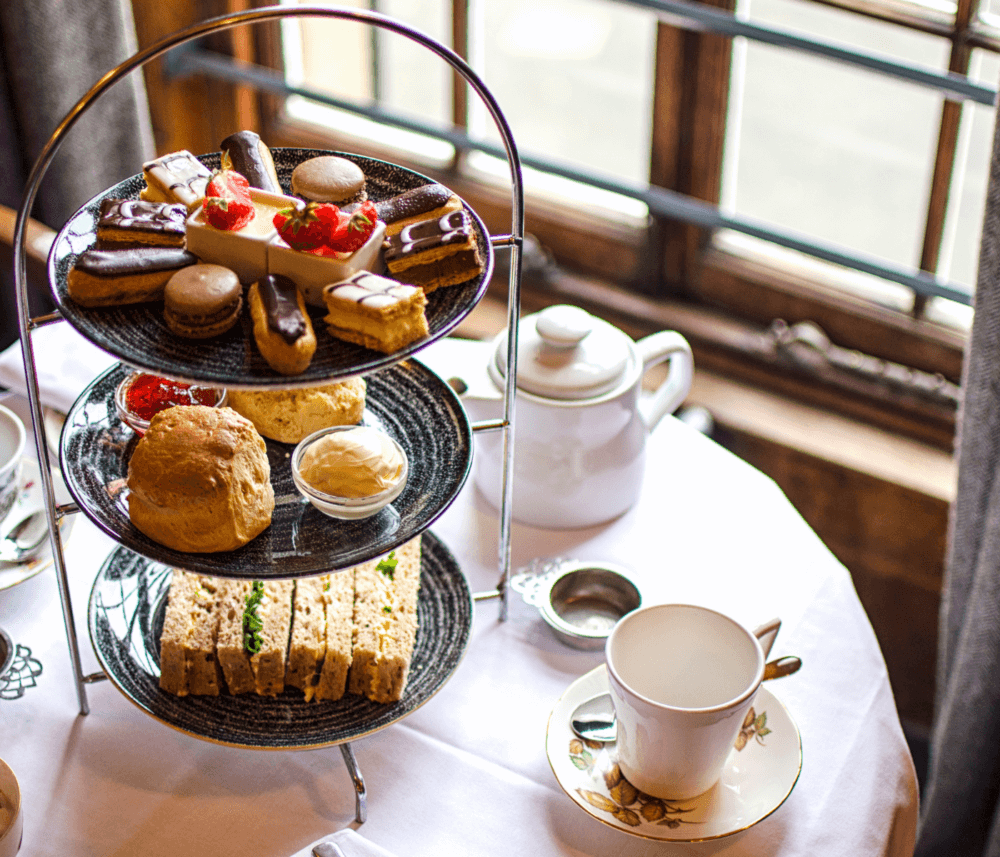 A three-tiered tray with assorted pastries, scones, sandwiches, and tea set on a table near a window. - Home Instead