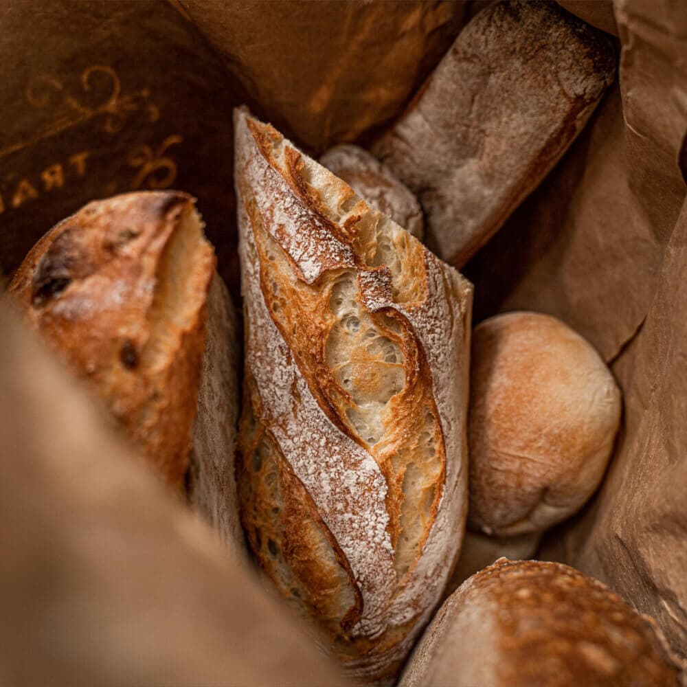 A variety of freshly baked bread loaves in a brown paper bag. - Home Instead
