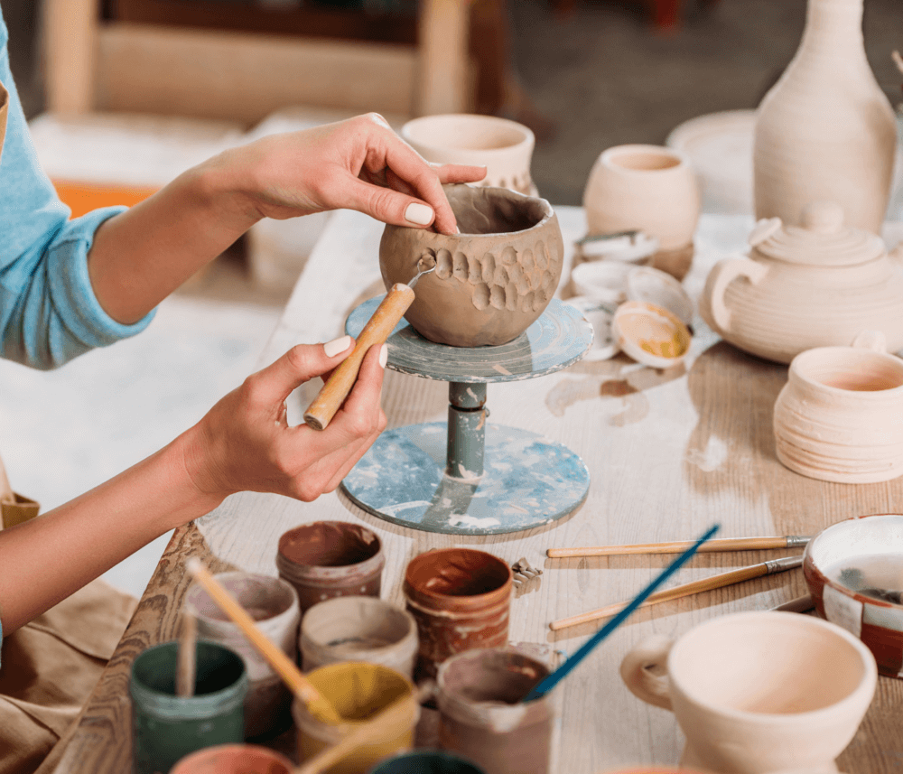 Person sculpting a clay pot with various pottery tools and paint cups on a wooden table. - Home Instead