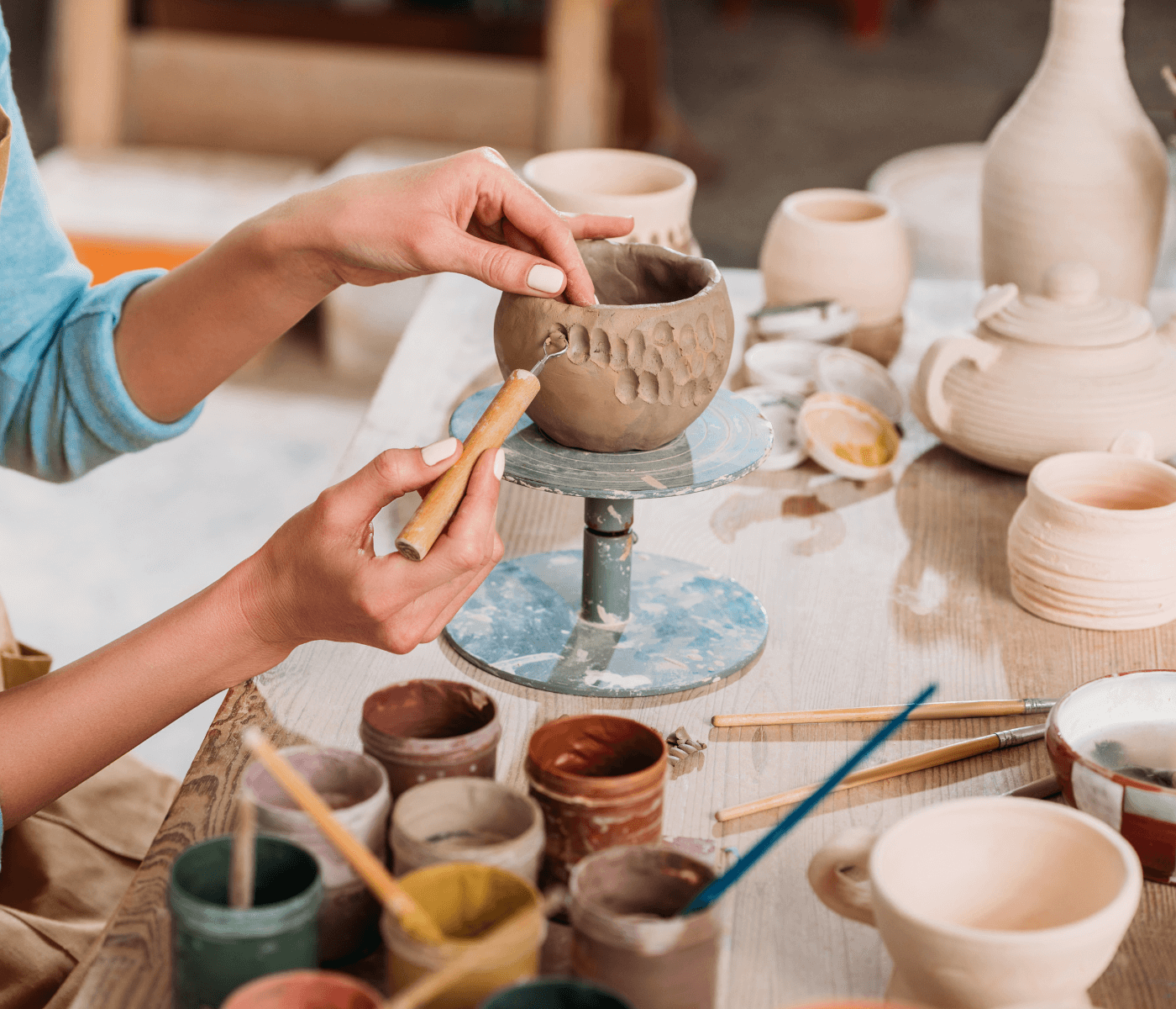Person sculpting a clay pot with various pottery tools and paint cups on a wooden table. - Home Instead