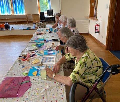 A group of elderly women sitting at a long table, engaging in arts and crafts activities in a room with blue curtains. - Home Instead