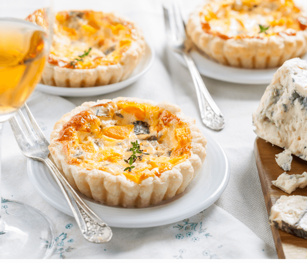Close-up of mini quiches with garnish, served on white plates. A glass of wine and a cheese block are beside the quiches. - Home Instead