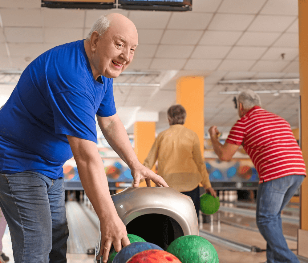 Elderly man in a blue shirt picking up a bowling ball with friends bowling in the background at a bowling alley. - Home Instead