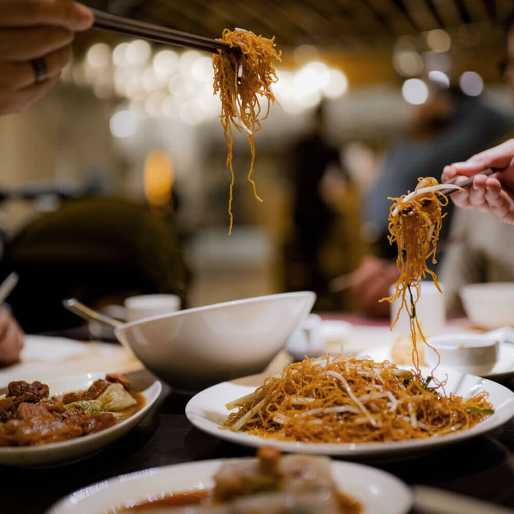 People using chopsticks to eat noodles from shared dishes on a dining table, with a blurred background. - Home Instead