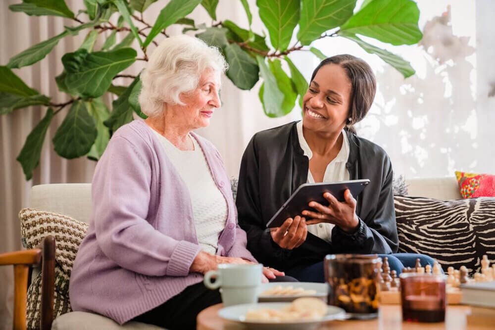 An elderly woman and a younger woman smiling at each other while looking at a tablet in a cozy living room. - Home Instead