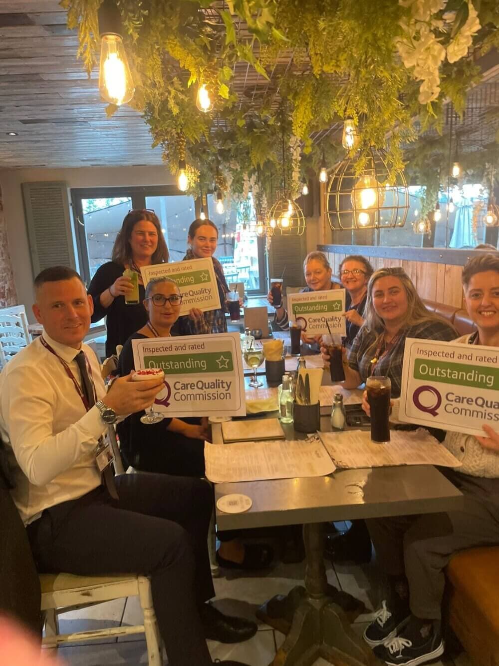 A group of people sit in a restaurant holding certificates with "Outstanding Care Quality Commission" written on them. - Home Instead