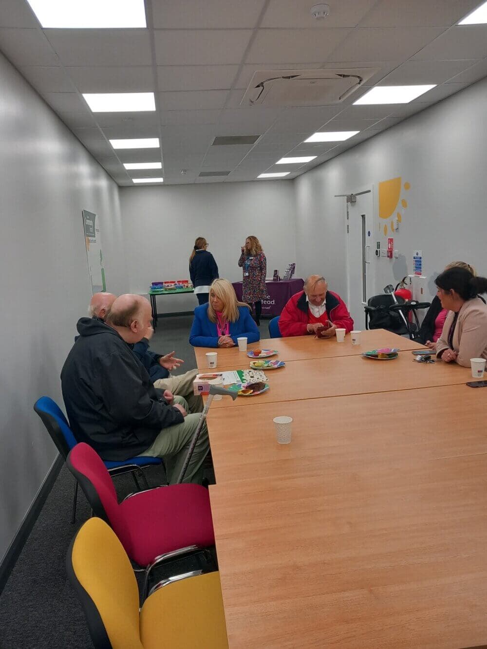 People sitting at a conference room table, talking and having snacks. A few others are in the background near a booth. - Home Instead