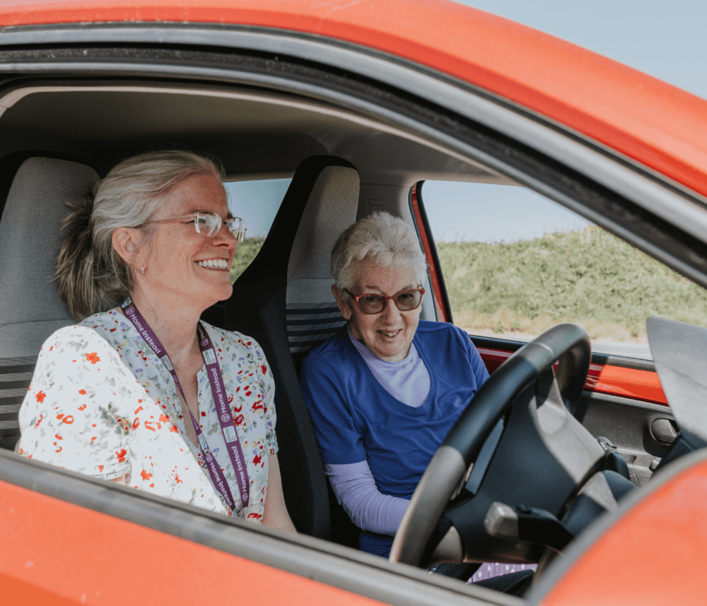 Two elderly women smiling in a red car; one is the driver and the other is in the passenger seat. - Home Instead