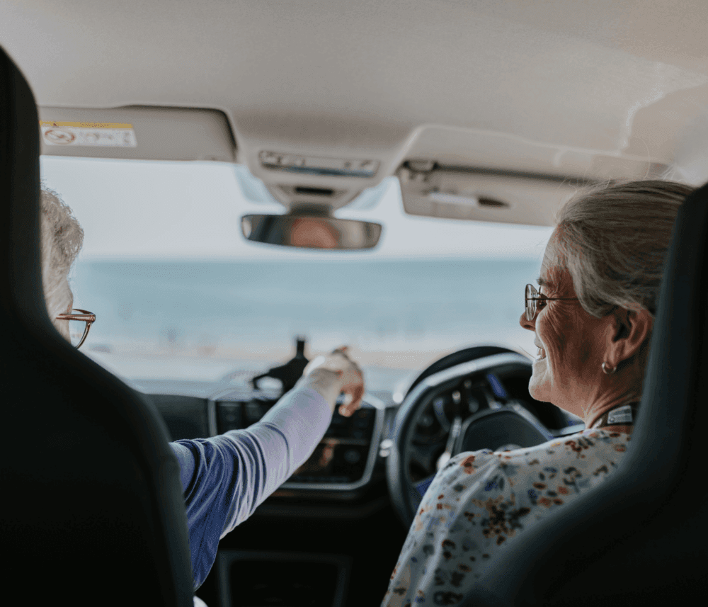 Two elderly women in a car, one driving and the other pointing at something outside, smiling and enjoying the view. - Home Instead