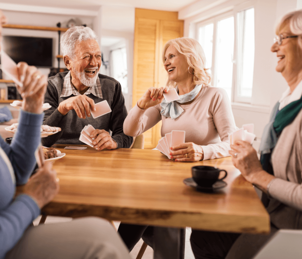 A group of elderly friends smiling and playing cards at a wooden table in a well-lit room. - Home Instead
