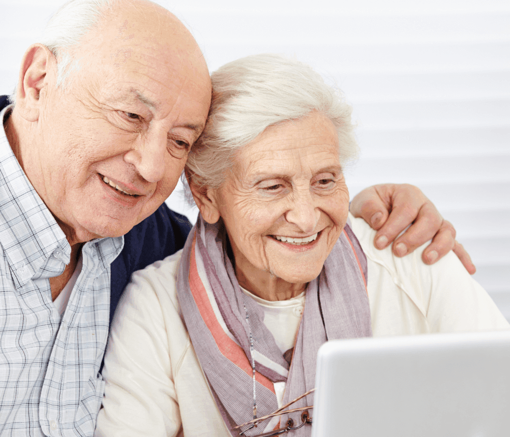 Elderly couple smiling while looking at a laptop screen together, with the man’s arm around the woman’s shoulders. - Home Instead