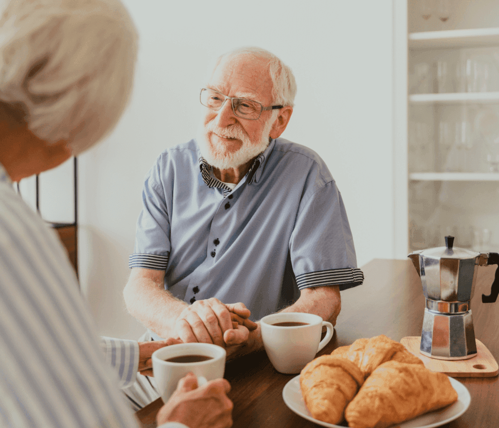 Two elderly people enjoying coffee and croissants at a table, sharing a warm conversation and holding hands. - Home Instead