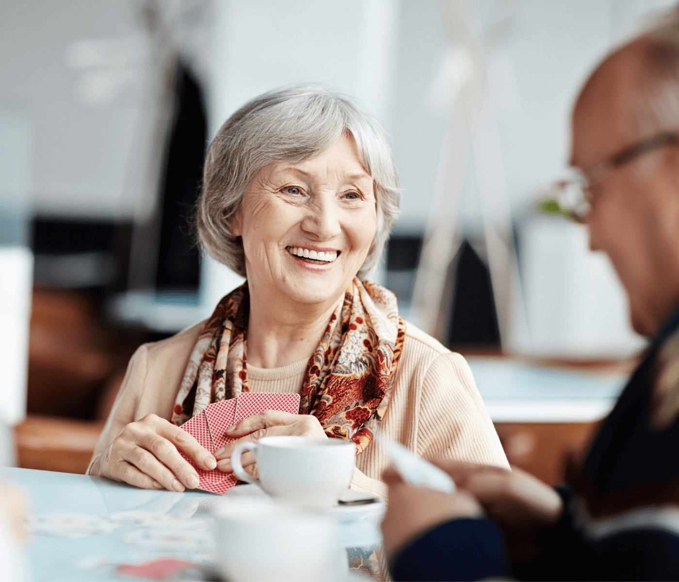 Smiling elderly woman holding playing cards, sitting at a table with a cup of coffee, conversing with a man. - Home Instead