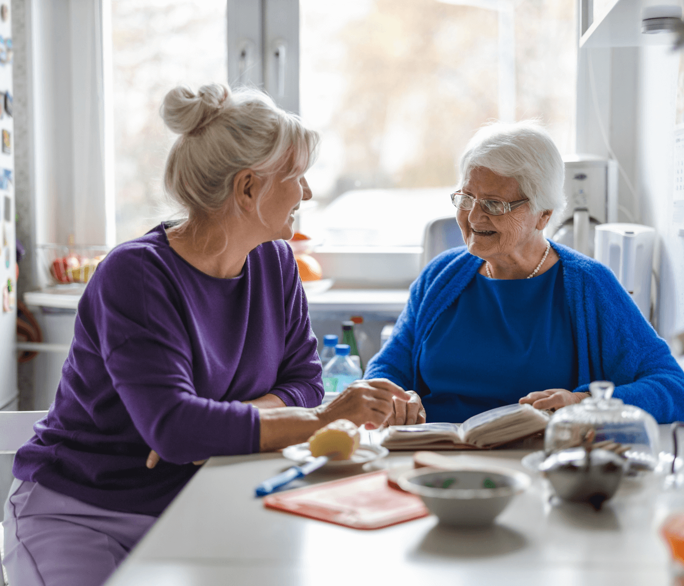 Two elderly women wearing glasses and casual clothes sit at a kitchen table, smiling, with books and food around them. - Home Instead