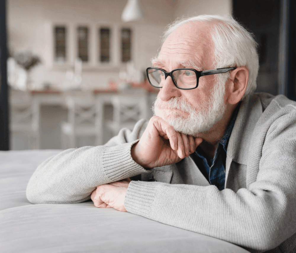 An elderly man with glasses and a gray beard looks out thoughtfully, resting his chin on his hand. - Home Instead