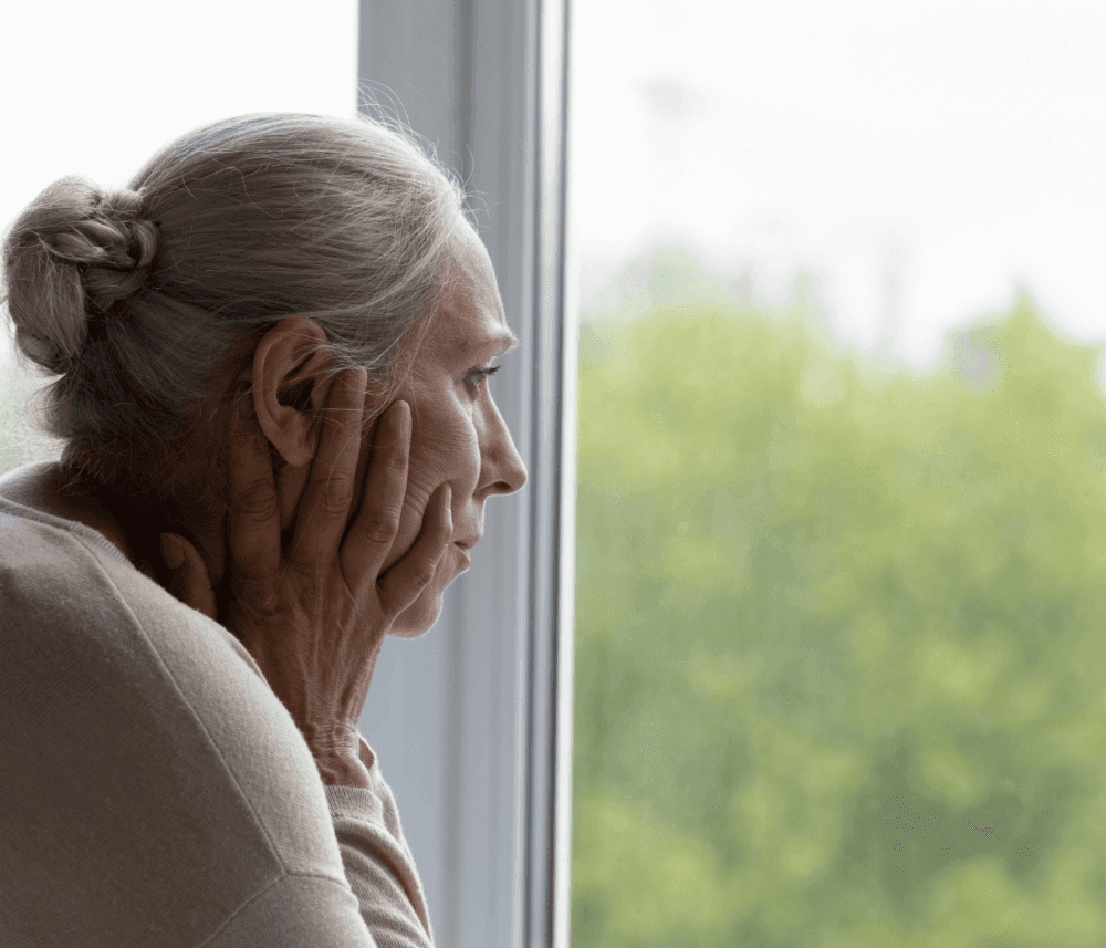 An elderly woman with gray hair looks pensively out of a window with her hands on her face. - Home Instead
