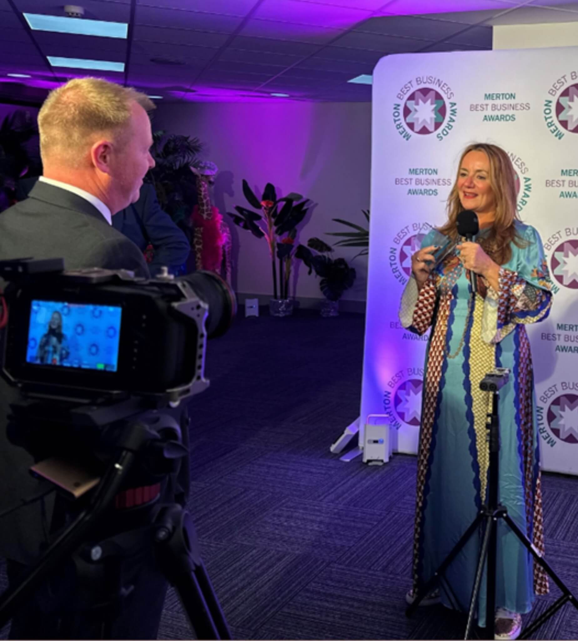 A woman in a colorful dress speaks on camera at the Merton Best Business Awards, with a cameraman filming her. - Home Instead