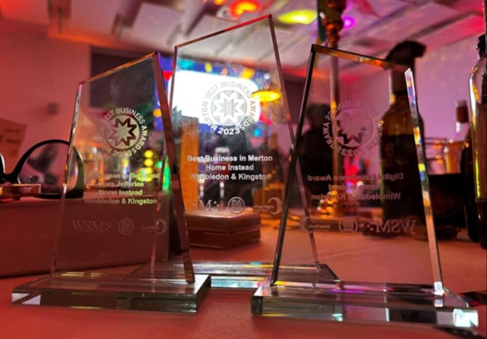 Three engraved glass business awards on a table with colorful lights in the background. - Home Instead