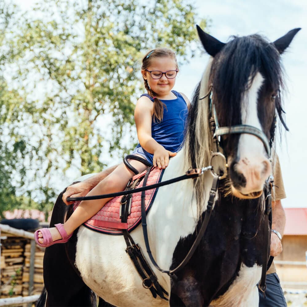 A girl with glasses rides a black and white horse on a sunny day, smiling and holding the reins. - Home Instead