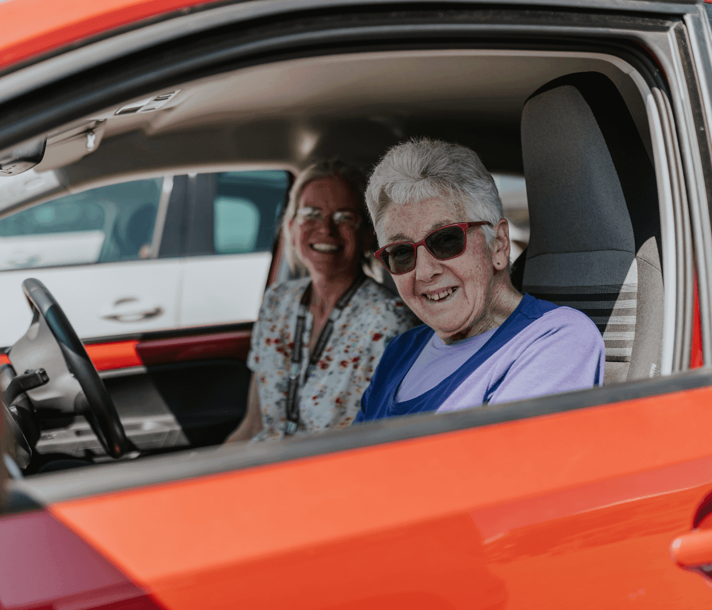 Two women smiling and sitting inside a red car, with one wearing sunglasses and the other in the background. - Home Instead