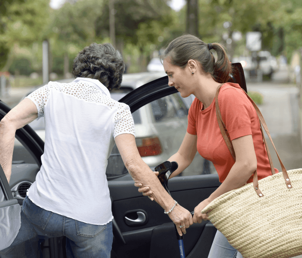 A woman helps an elderly person with a cane get into a car, holding the door open on a street with trees in the background. - Home Instead