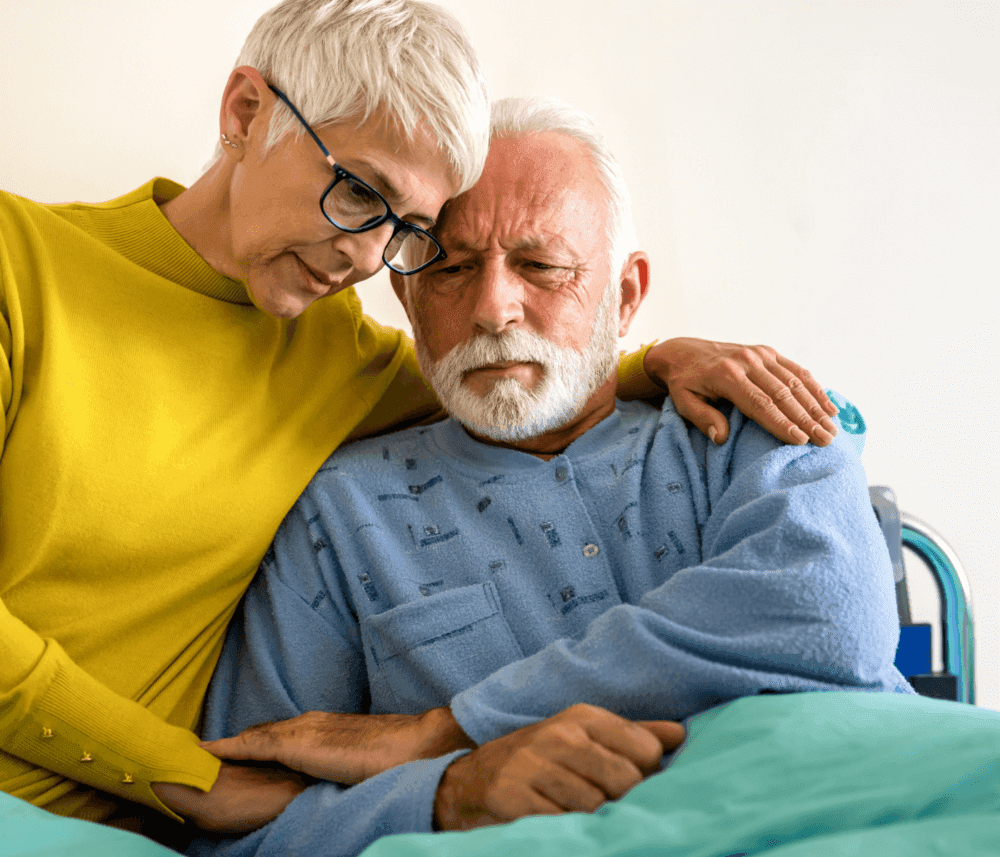 Woman with short gray hair in yellow sweater comforting elderly man with white beard in light blue clothing on a hospital bed. - Home Instead